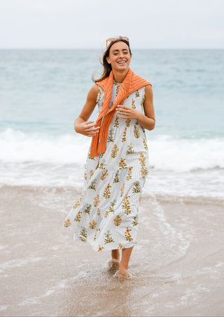 Woman in a floral dress and orange scarf standing on a beach with ocean waves in the background